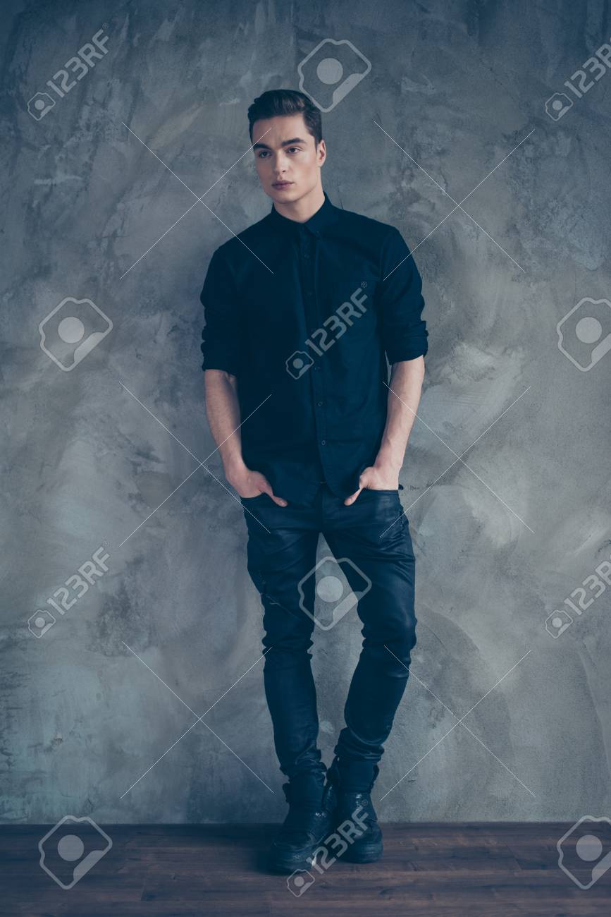 Modelo Atractivo Joven En Camisa Negra, Jeans Y Elegantes Botas De Pie Sobre Fondo Gris, Se Metió Las Manos En Bolsillos. Fotos, Retratos, Imágenes Y Fotografía De Archivo Libres De Derecho.