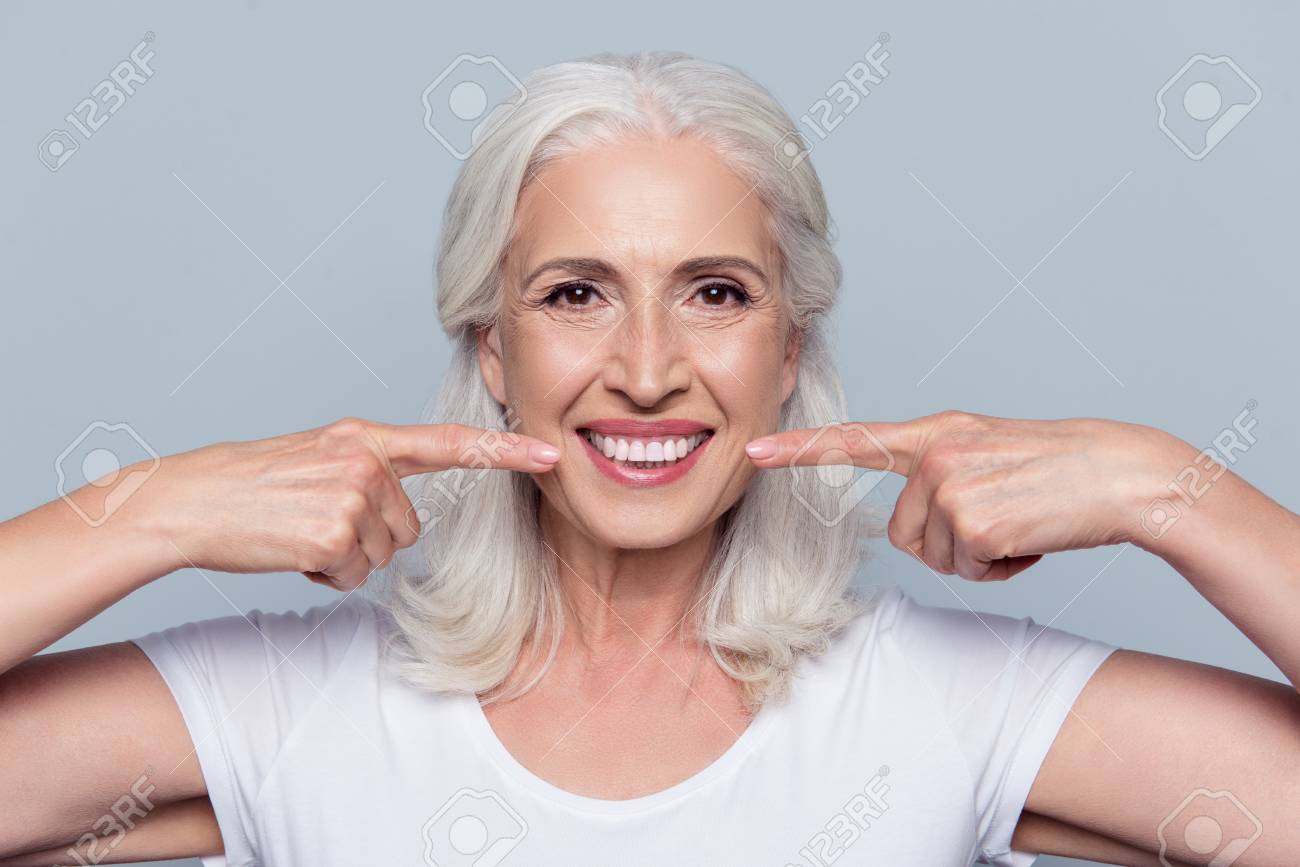 Concept of having strong healthy straight white teeth at old age. Close up portrait of happy with beaming smile female pensioner pointing on her perfect clear white teeth, isolated on gray background - 91651657 Concept of having strong healthy straight white teeth at old age. Close up portrait of happy with beaming smile female pensioner pointing on her perfect clear white teeth, isolated on gray background - 91651657