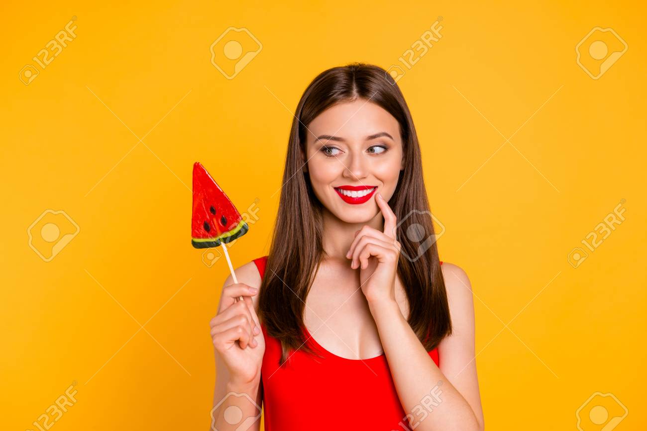 To be or not to be? Close up portrait happy and cute young woman holding piece on caramel fruit on stick, looking thoughtfully aside isolated on bright yellow background - 106119554 To be or not to be? Close up portrait happy and cute young woman holding piece on caramel fruit on stick, looking thoughtfully aside isolated on bright yellow background - 106119554