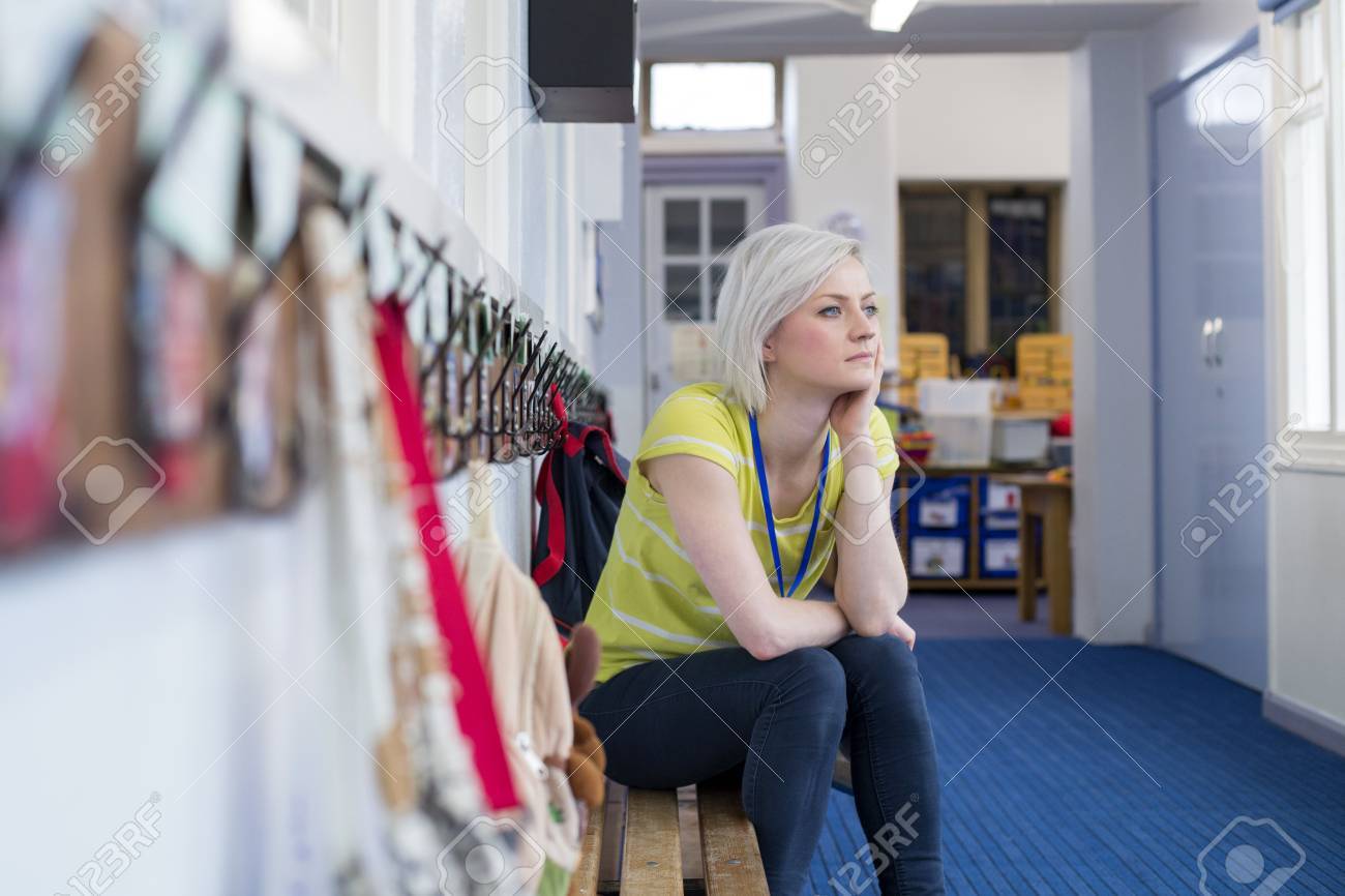 Young, female teacher sitting on a bench in the cloakroom of school. She is looking pensively out of the window. - 60255969 Young, female teacher sitting on a bench in the cloakroom of school. She is looking pensively out of the window. - 60255969