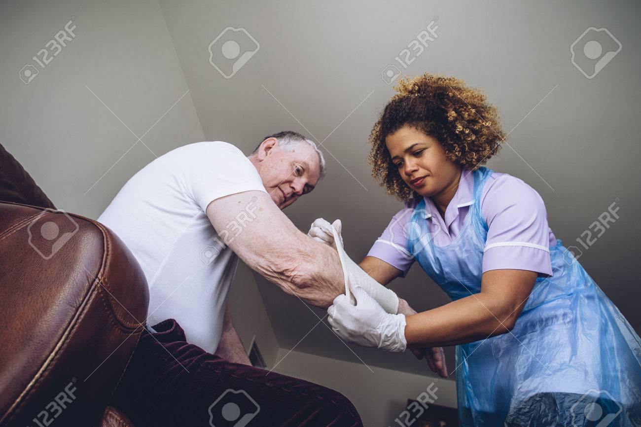 Caregiving Nurse putting a bandage on a senior mans arm in his home. - 65447309 Caregiving Nurse putting a bandage on a senior mans arm in his home. - 65447309
