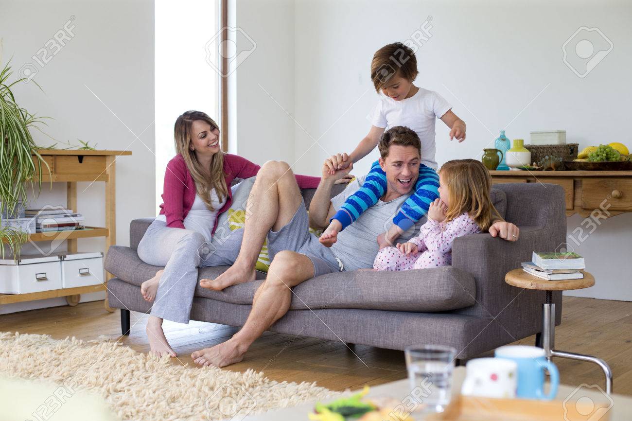 Family of four are sitting on the sofa in the living room of their home together. The father has his son on his shoulders and the little girl is sitting next to him. - 68981205 Family of four are sitting on the sofa in the living room of their home together. The father has his son on his shoulders and the little girl is sitting next to him. - 68981205