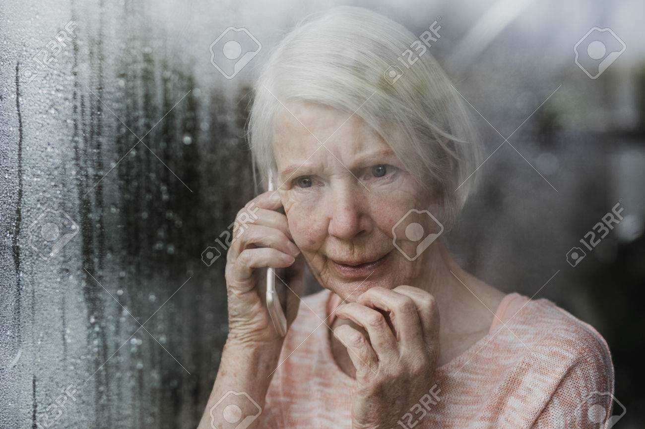 Senior woman is looking worriedly out of the window of her home while talking to someone on the phone. - 85444214 Senior woman is looking worriedly out of the window of her home while talking to someone on the phone. - 85444214