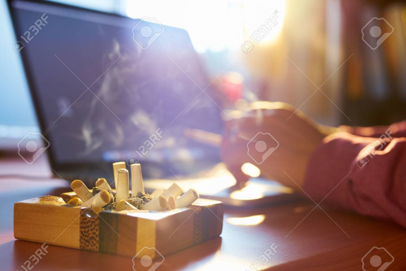 Close up of ashtray full of cigarette, with man in background working on laptop computer and smoking indoors on early morning. Concept of addiction and abuse of nicotine. - 42016233 Close up of ashtray full of cigarette, with man in background working on laptop computer and smoking indoors on early morning. Concept of addiction and abuse of nicotine. - 42016233