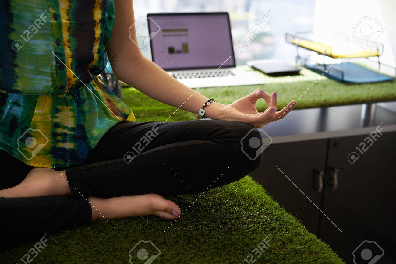Young hispanic woman in office, sitting on desk covered with grass and plants. She meditates yoga in lotus position. Cropped view of hands and feet - 51500359 Young hispanic woman in office, sitting on desk covered with grass and plants. She meditates yoga in lotus position. Cropped view of hands and feet - 51500359
