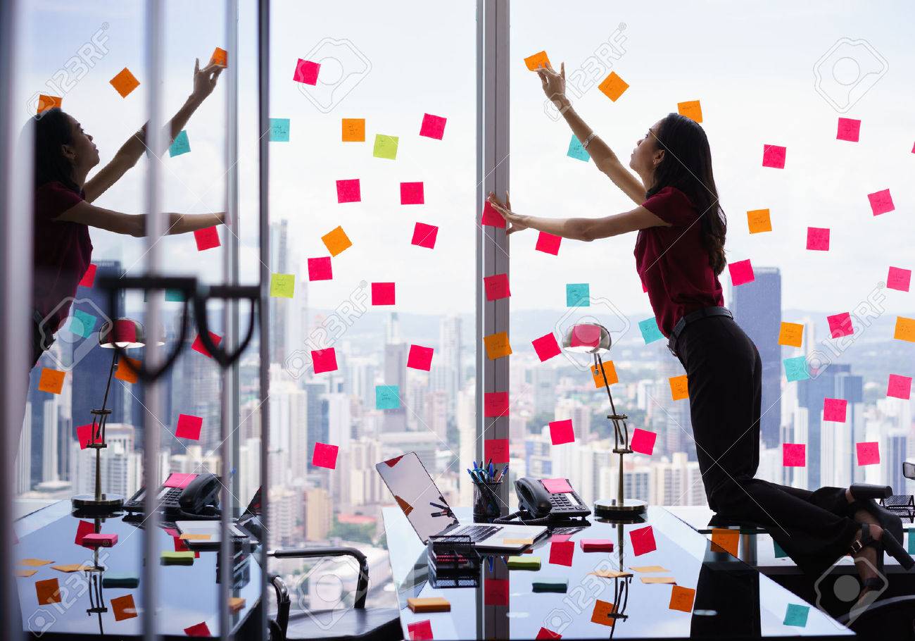 Mixed race secretary working in modern office in skyscraper, sticking adhesive notes with tasks on window. The girl feels stressed and overwhelmed - 51500374 Mixed race secretary working in modern office in skyscraper, sticking adhesive notes with tasks on window. The girl feels stressed and overwhelmed - 51500374