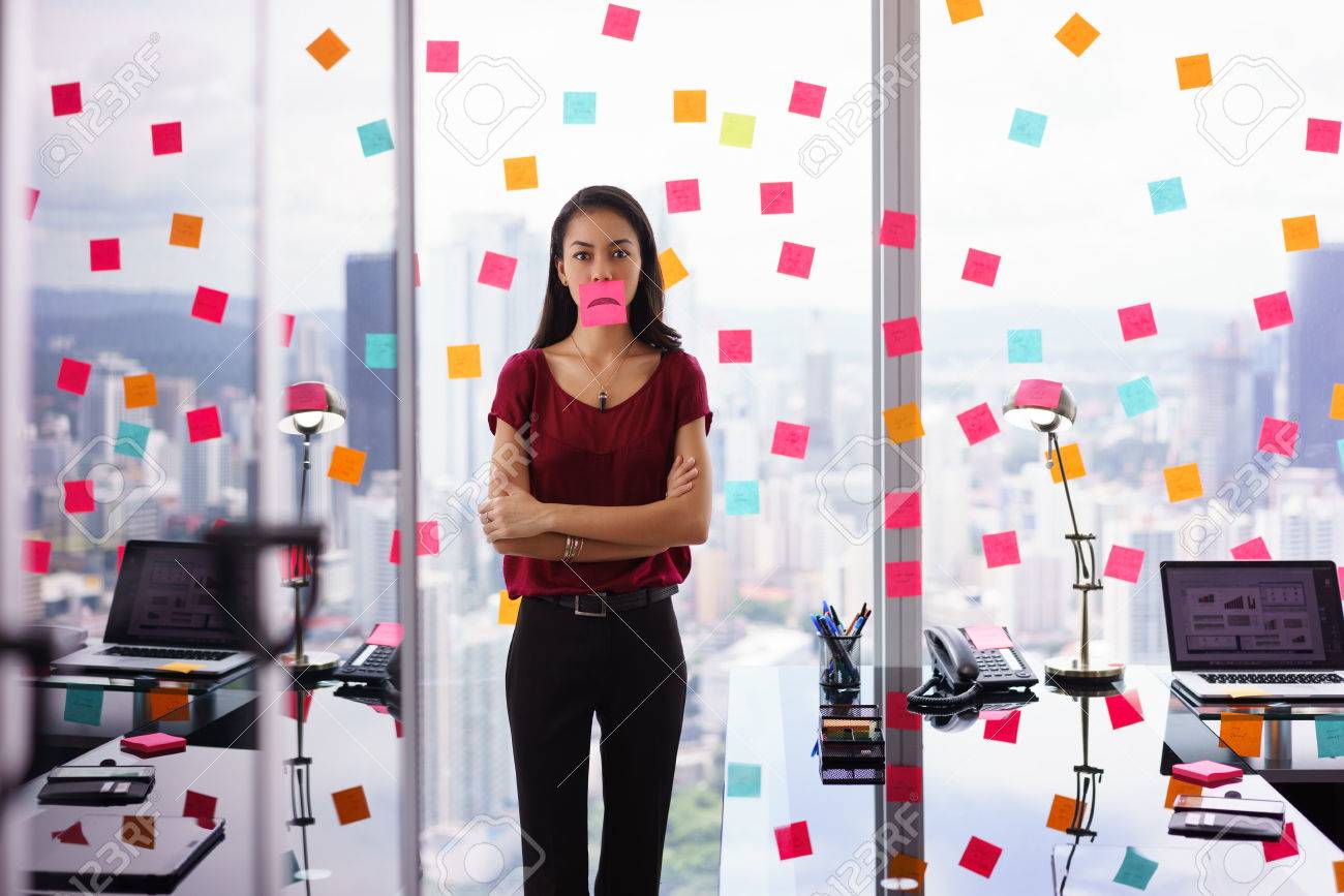 Mixed race woman working in modern office with reminders on skyscraper window. The girl feels stressed, holds a sticking note with sad emoticon on mouth. - 51500377 Mixed race woman working in modern office with reminders on skyscraper window. The girl feels stressed, holds a sticking note with sad emoticon on mouth. - 51500377