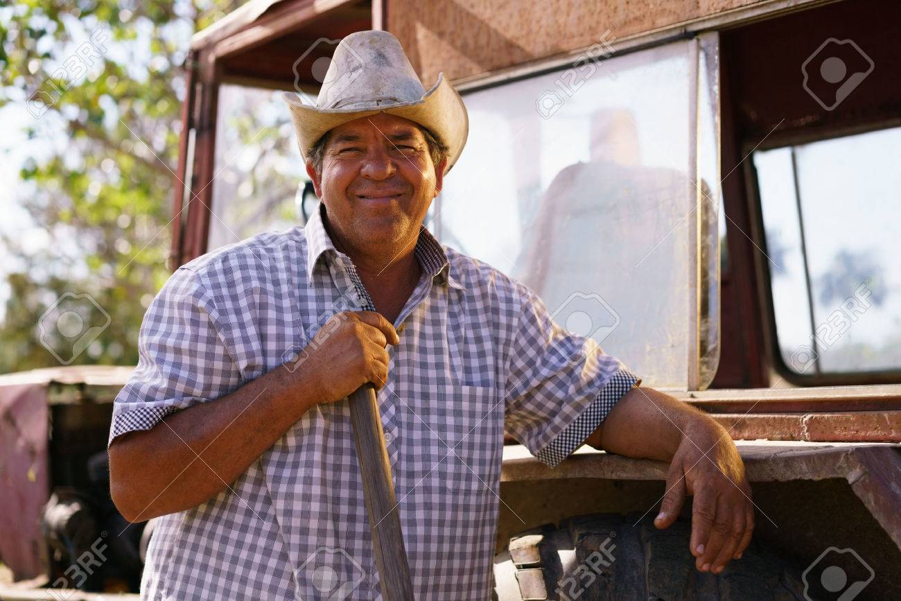 Farming and cultivations in Latin America. Portrait of middle aged hispanic farmer sitting proud in his tractor at sunset, holding the volante. He looks at the camera and smiles happy. - 56140488 Farming and cultivations in Latin America. Portrait of middle aged hispanic farmer sitting proud in his tractor at sunset, holding the volante. He looks at the camera and smiles happy. - 56140488