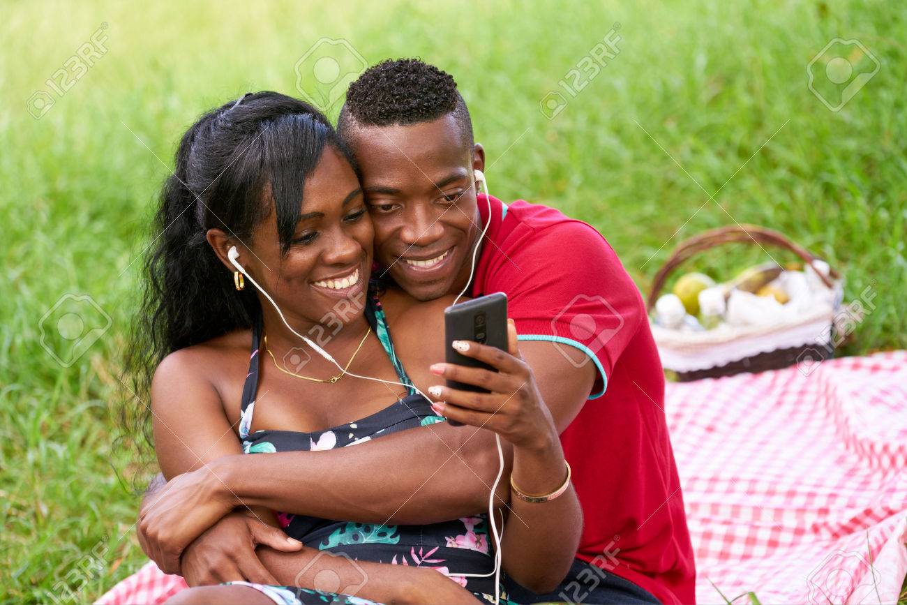 都市公園で楽しんで幸せな黒のカップル 若いアフリカ系アメリカ人男性と女性音楽を聞くと携帯電話で動画を見てします の写真素材 画像素材 Image