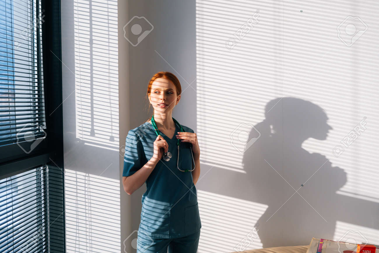 Close-up of unrecognizable female doctor in blue green uniform putting on stethoscope on neck standing near window in sunny day. Closeup of woman physician wearing stethoscope. - 169892793 Close-up of unrecognizable female doctor in blue green uniform putting on stethoscope on neck standing near window in sunny day. Closeup of woman physician wearing stethoscope. - 169892793