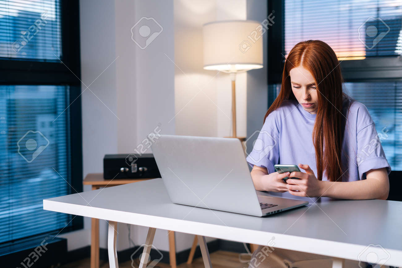 Serious young woman using phone and sitting at desk with laptop computer in apartment near window evening at late. Attractive redhead lady working from home office looking at screen of smartphone. - 169892796 Serious young woman using phone and sitting at desk with laptop computer in apartment near window evening at late. Attractive redhead lady working from home office looking at screen of smartphone. - 169892796
