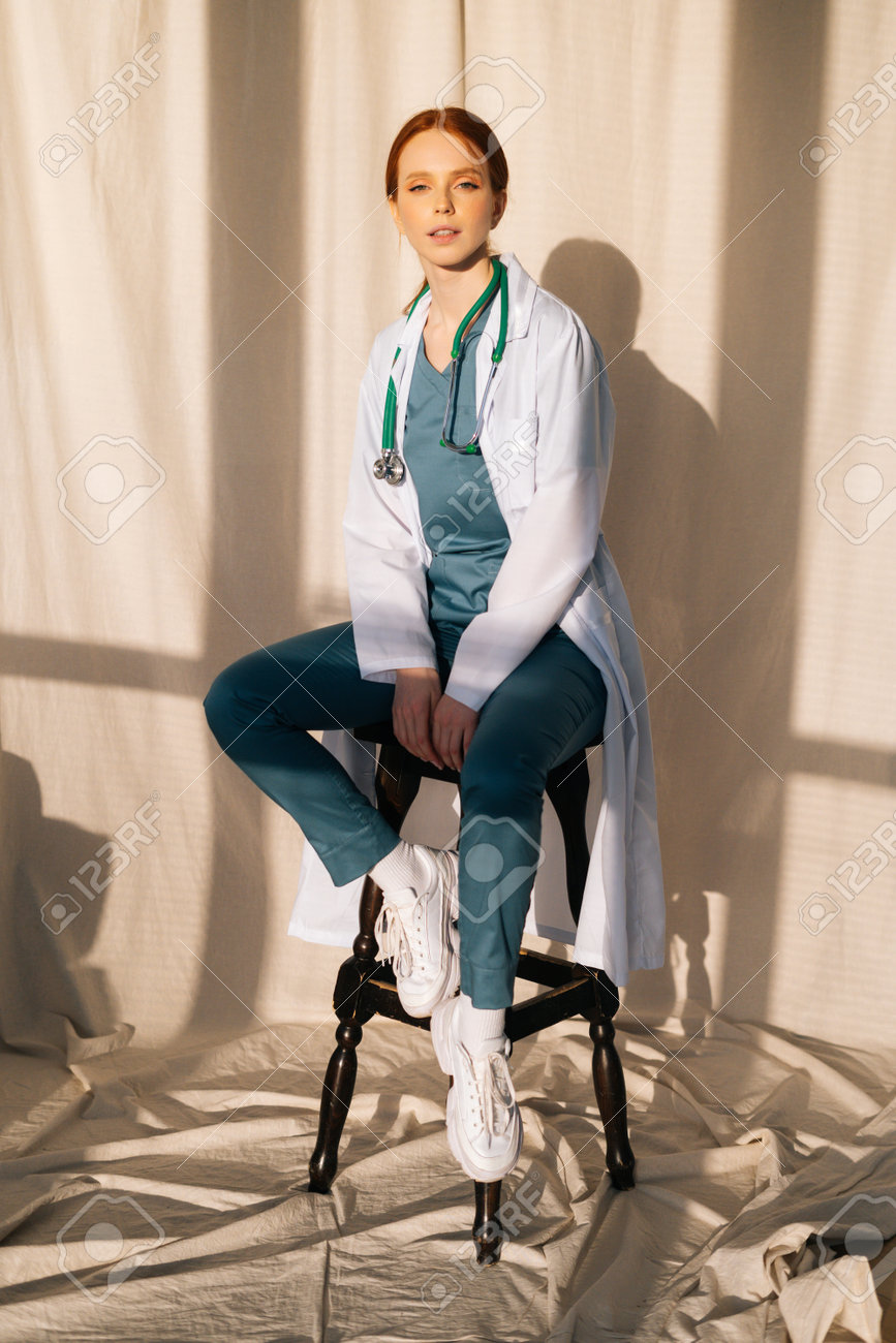 Portrait of serious sensual female doctor in white coat sitting on chair near window in sunny day in medical clinic office. Young redhead woman surgeon posing with stethoscope looking at camera. - 169892799 Portrait of serious sensual female doctor in white coat sitting on chair near window in sunny day in medical clinic office. Young redhead woman surgeon posing with stethoscope looking at camera. - 169892799