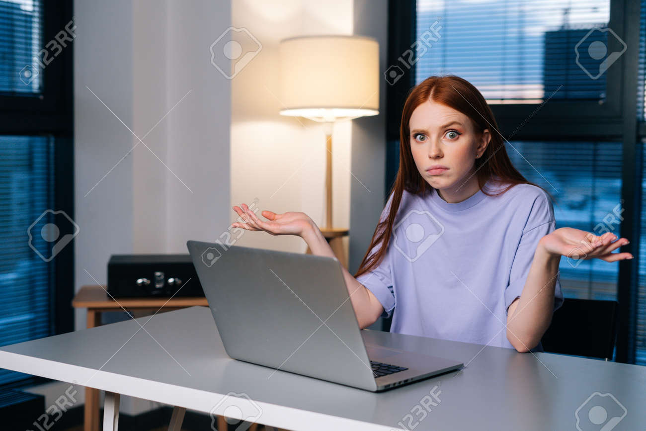 Frustrated young woman confused by laptop computer problem sitting at desk in home office near window evening at late, looking at camera. Bewildered female spreads hands incomprehensibly. - 169892805 Frustrated young woman confused by laptop computer problem sitting at desk in home office near window evening at late, looking at camera. Bewildered female spreads hands incomprehensibly. - 169892805