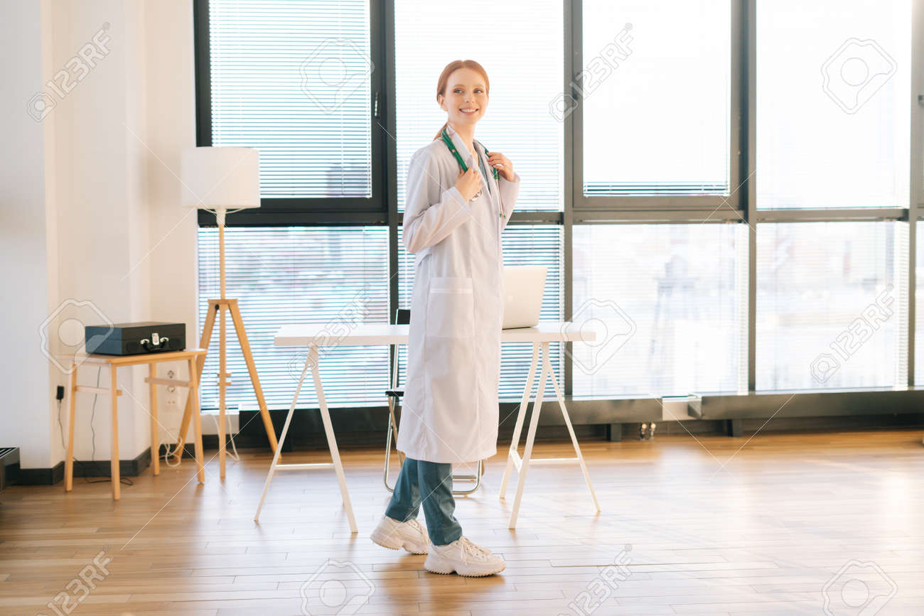 Front view of charming female doctor in white coat standing on background of window in sunny day in light medical clinic office. Young redhead woman physician posing with stethoscope looking away. - 169892815 Front view of charming female doctor in white coat standing on background of window in sunny day in light medical clinic office. Young redhead woman physician posing with stethoscope looking away. - 169892815