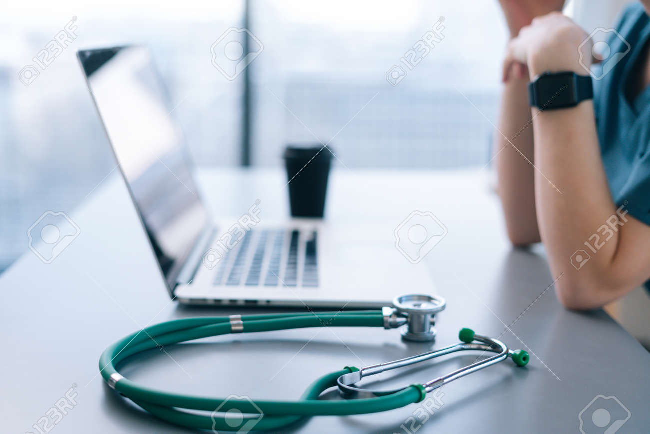 Side view of thoughtful unrecognizable female practitioner doctor sitting at desk with laptop sitting in hospital office of medic clinic, stethoscope and coffee cup on table. - 169892818 Side view of thoughtful unrecognizable female practitioner doctor sitting at desk with laptop sitting in hospital office of medic clinic, stethoscope and coffee cup on table. - 169892818