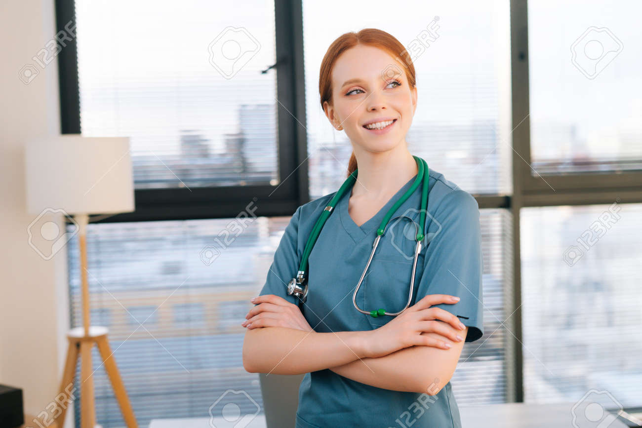 Portrait of happy female doctor in blue green uniform standing arms crossed near window in sunny day in medical clinic office. Redhead woman surgeon posing with stethoscope looking away. - 169892819 Portrait of happy female doctor in blue green uniform standing arms crossed near window in sunny day in medical clinic office. Redhead woman surgeon posing with stethoscope looking away. - 169892819
