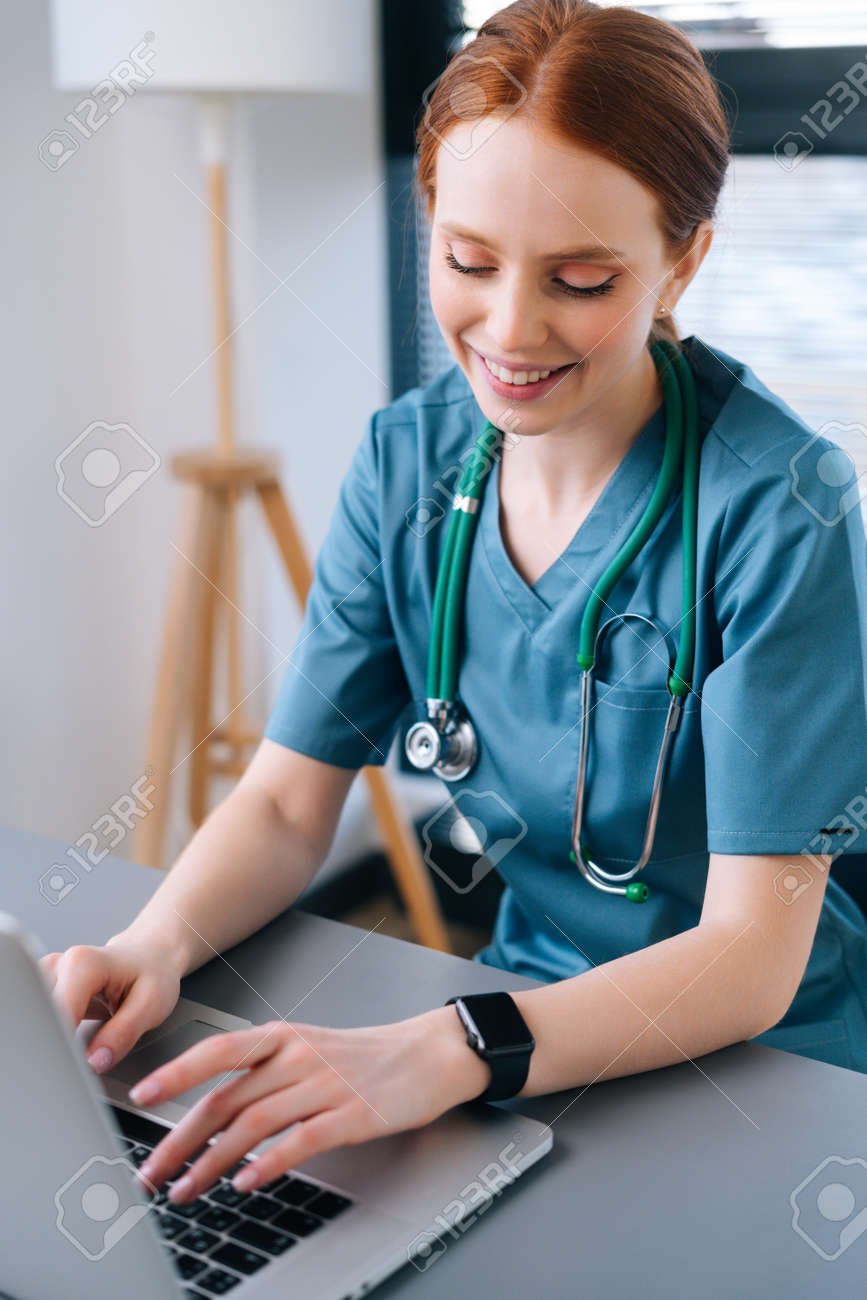 Close-up of pretty smiling young female doctor in blue green medical uniform sitting at desk with laptop on background of window in hospital office of medic clinic, looking down. - 169892826 Close-up of pretty smiling young female doctor in blue green medical uniform sitting at desk with laptop on background of window in hospital office of medic clinic, looking down. - 169892826
