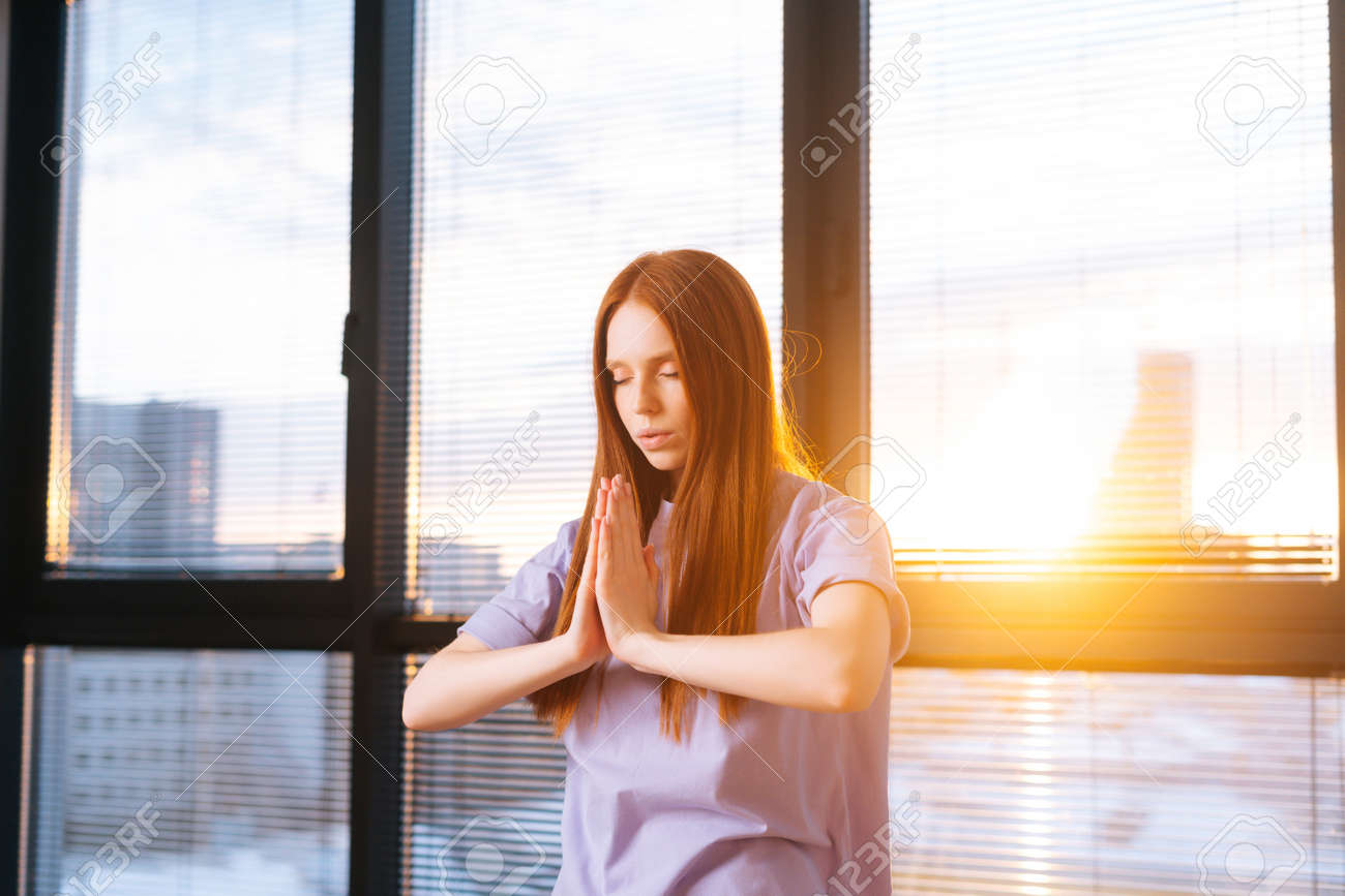 Close-up of peaceful redhead young woman practicing yoga holding hands in namaste mudra posture, standing near large window at sunset. Yogi female putting hands in namaste posture and relaxing. - 169892829 Close-up of peaceful redhead young woman practicing yoga holding hands in namaste mudra posture, standing near large window at sunset. Yogi female putting hands in namaste posture and relaxing. - 169892829
