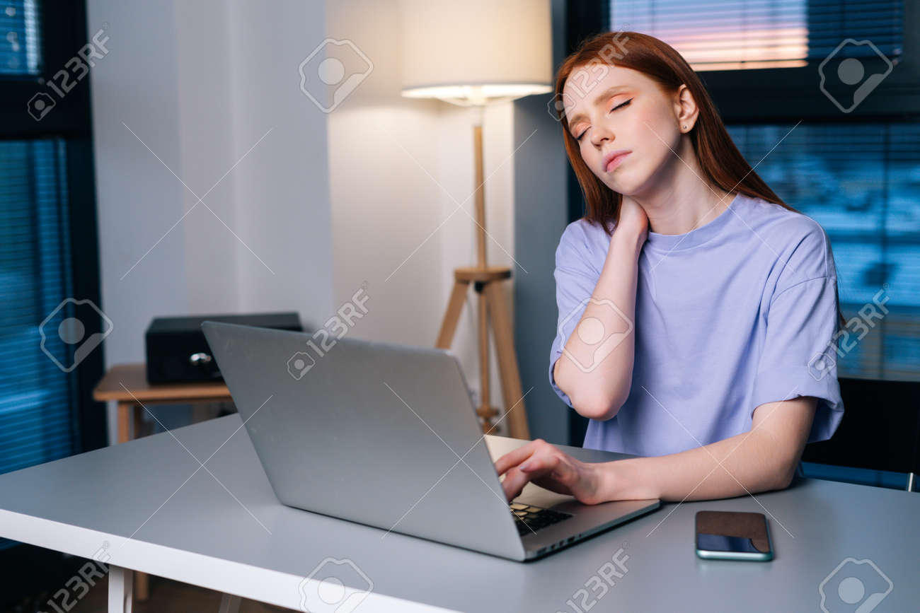 Overworked redhead young woman typing on laptop actively feeling pain in wrist sitting at desk near window in evening. Lady working on laptop and suffering from pain in hand, carpal tunnel syndrome. - 169892832 Overworked redhead young woman typing on laptop actively feeling pain in wrist sitting at desk near window in evening. Lady working on laptop and suffering from pain in hand, carpal tunnel syndrome. - 169892832