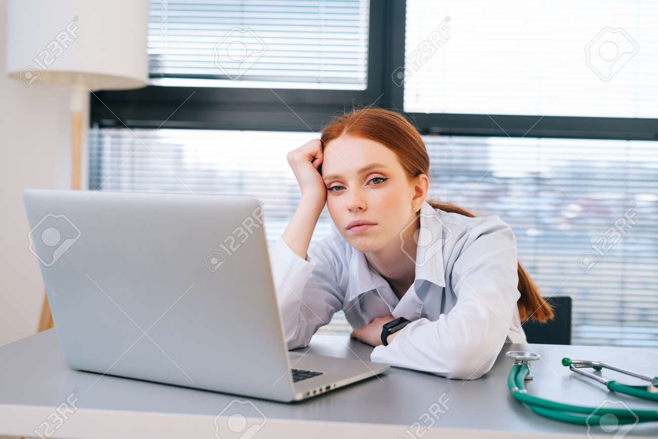 Close-up portrait of tired exhausted young female doctor in white coat sitting at desk with laptop. Front view of overworked woman physician in hospital office looking at camera. - 169892861 Close-up portrait of tired exhausted young female doctor in white coat sitting at desk with laptop. Front view of overworked woman physician in hospital office looking at camera. - 169892861
