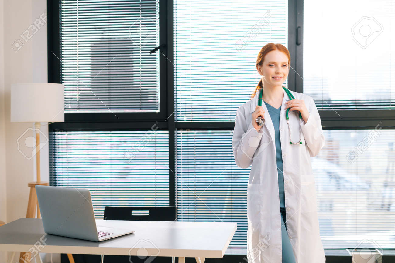 Portrait of pretty female doctor in white coat standing on background of window in sunny day in light medical clinic office. Young redhead woman physician posing with stethoscope looking at camera. - 169892865 Portrait of pretty female doctor in white coat standing on background of window in sunny day in light medical clinic office. Young redhead woman physician posing with stethoscope looking at camera. - 169892865