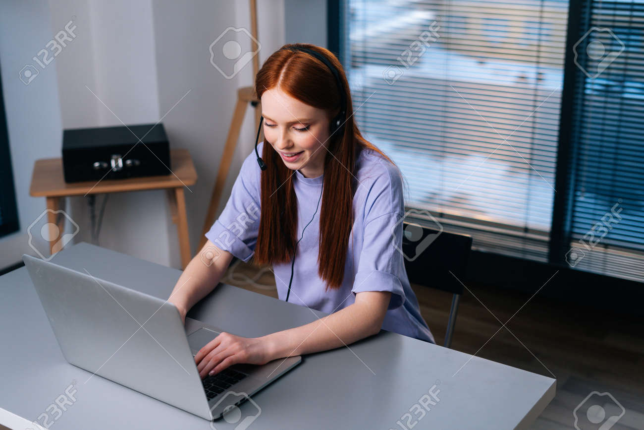Top view of attractive young woman operator using headset and laptop during customer support at home office. Young redhead female student communicating online by video call on background of window. - 169892870 Top view of attractive young woman operator using headset and laptop during customer support at home office. Young redhead female student communicating online by video call on background of window. - 169892870