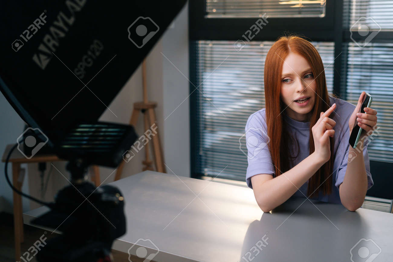 Webcam view of smiling young woman blogger greeting followers and talking about phone during video chat on background of window. POV of cheerful redhead female having presentation during sundown. - 169892872 Webcam view of smiling young woman blogger greeting followers and talking about phone during video chat on background of window. POV of cheerful redhead female having presentation during sundown. - 169892872
