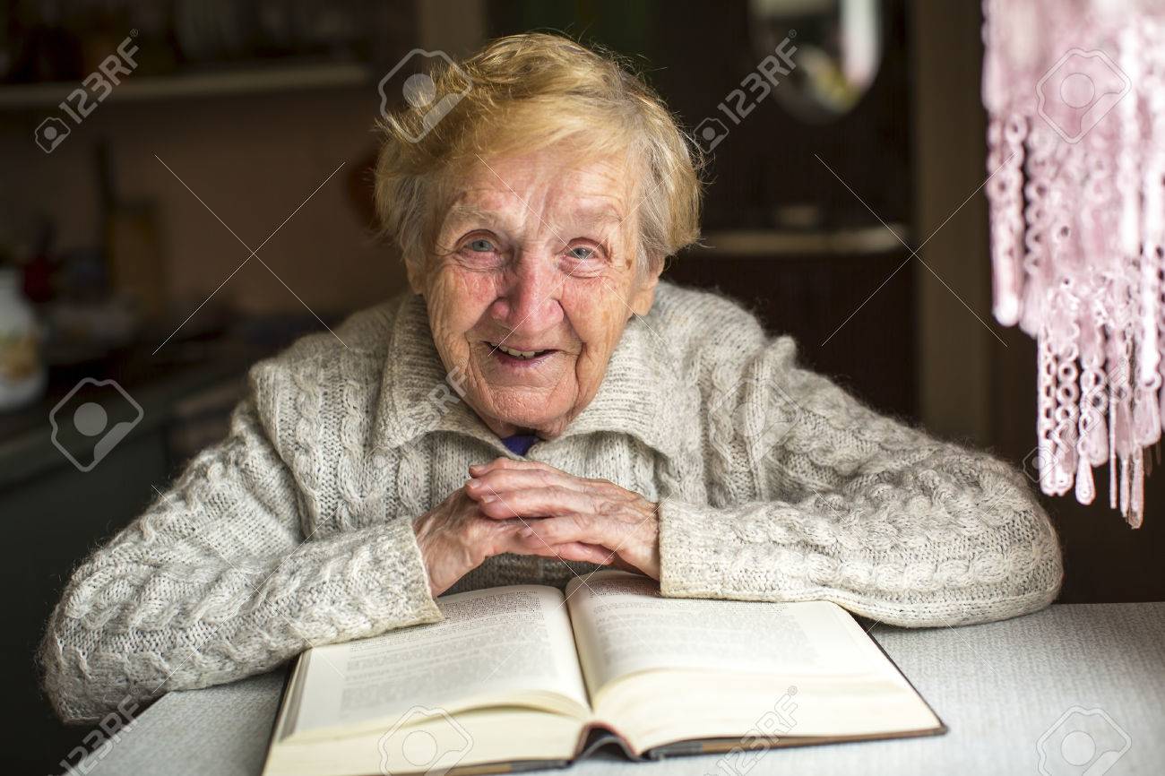 An elderly woman sits with a book at the table. - 48954949 An elderly woman sits with a book at the table. - 48954949