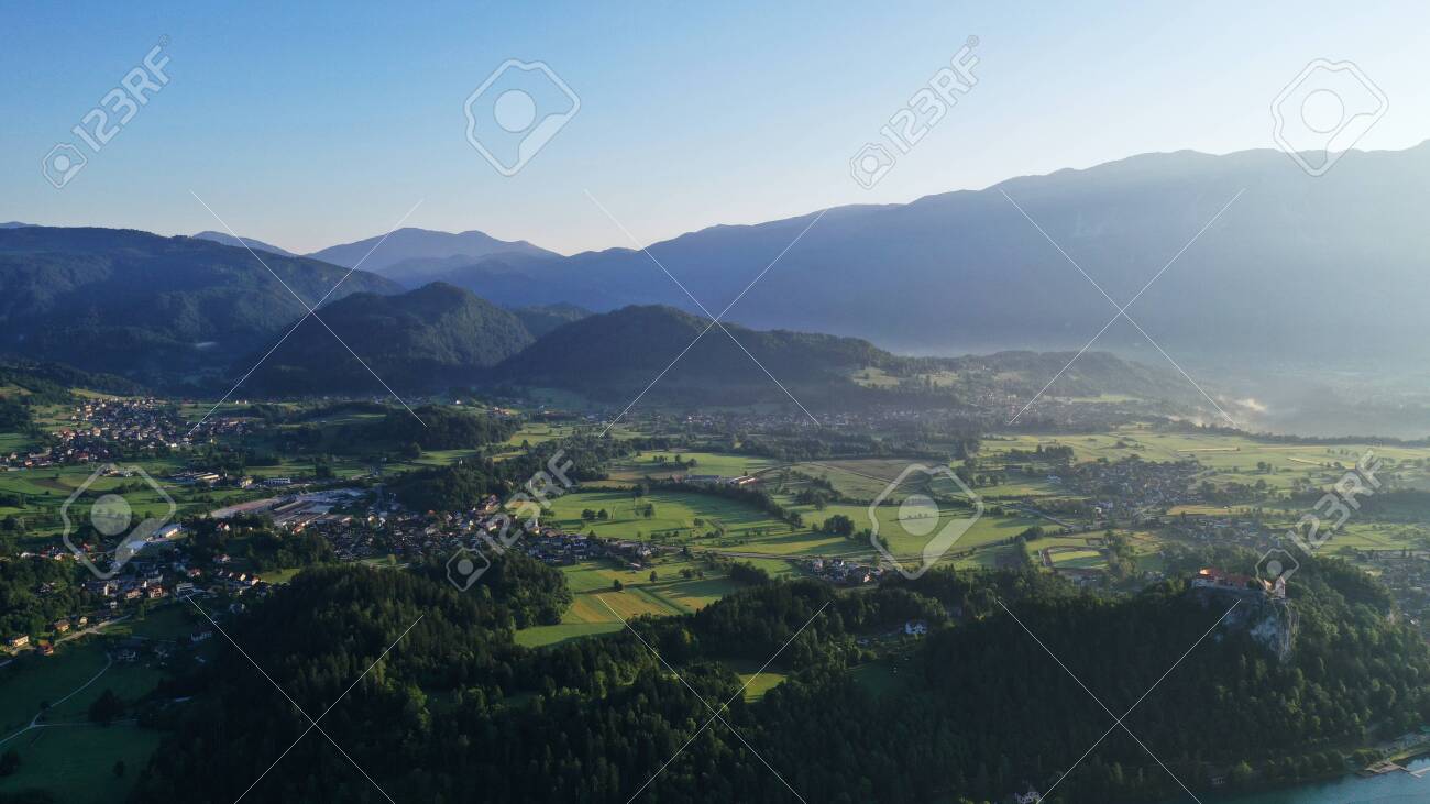 Aerial view of Bled Lake valley. Natural landscape with green meadows, fields, hills and mountains, small houses and trees. Sunny blue sky. Sun rays. - 139935514 Aerial view of Bled Lake valley. Natural landscape with green meadows, fields, hills and mountains, small houses and trees. Sunny blue sky. Sun rays. - 139935514