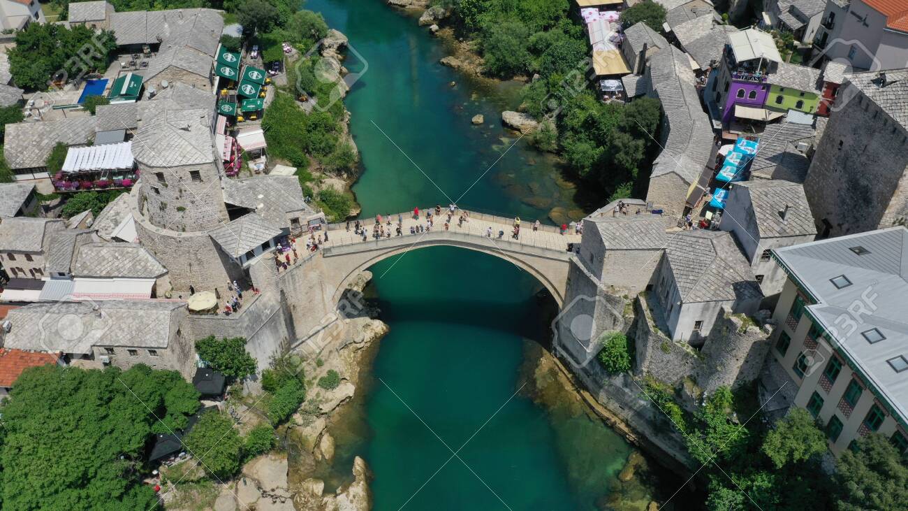Aerial view of Stari Most old medieval bridge in Mostar, Neretva river, Bosnia and Herzegovina. Tourists walking on the bridge. Summer landscape of old town. Mosque on background. - 139967243 Aerial view of Stari Most old medieval bridge in Mostar, Neretva river, Bosnia and Herzegovina. Tourists walking on the bridge. Summer landscape of old town. Mosque on background. - 139967243