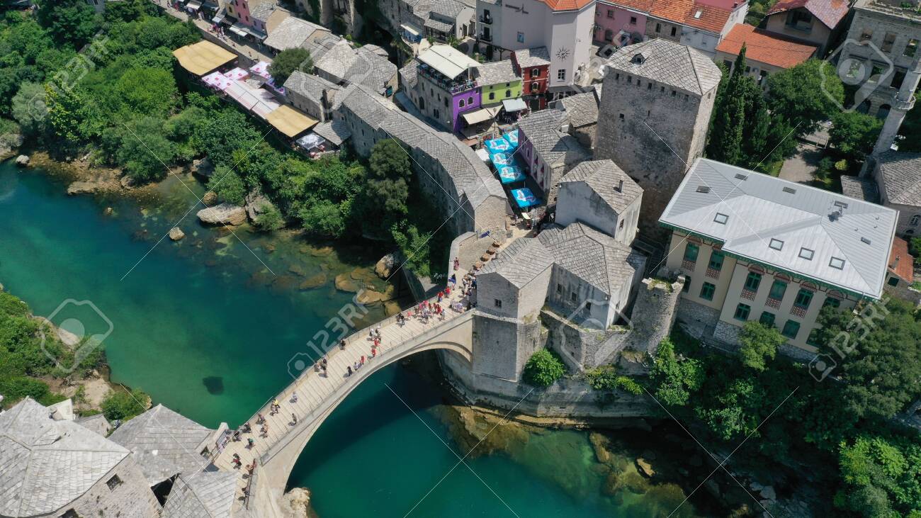 Aerial view of Stari Most old medieval bridge in Mostar, Neretva river, Bosnia and Herzegovina. Tourists walking on the bridge. Summer landscape of old town. Mosque on background. - 139967249 Aerial view of Stari Most old medieval bridge in Mostar, Neretva river, Bosnia and Herzegovina. Tourists walking on the bridge. Summer landscape of old town. Mosque on background. - 139967249
