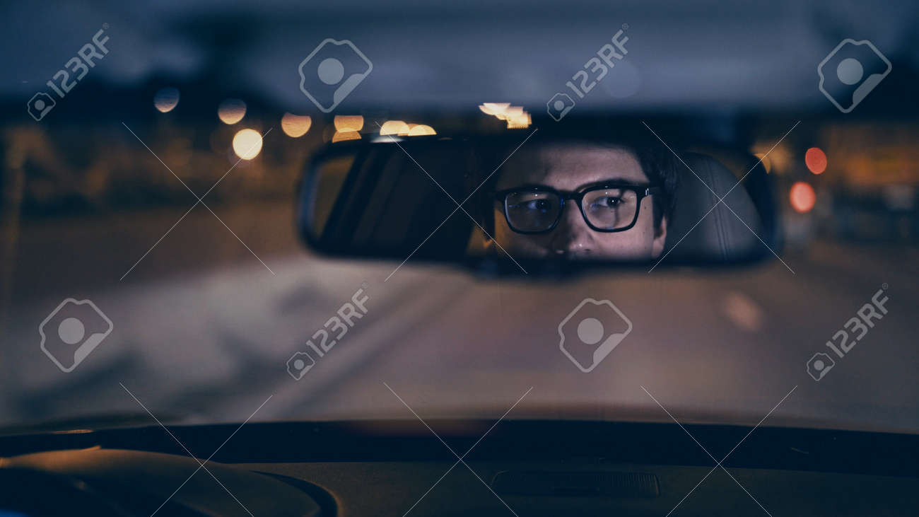 Drivers face is reflected in a rear view mirror, night shot, violet color, bokeh light. Tired concentrated driver in eyeglasses is reflected in a mirror in car, night road shot driving all night - 155325936 Drivers face is reflected in a rear view mirror, night shot, violet color, bokeh light. Tired concentrated driver in eyeglasses is reflected in a mirror in car, night road shot driving all night - 155325936