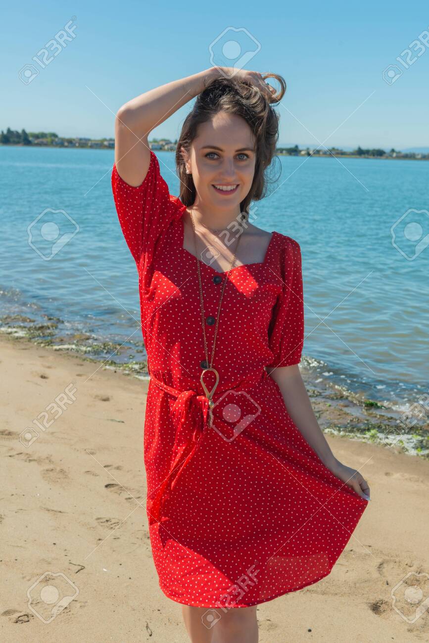 Petite Young Brunette In A Red Dress Standing On A Beach