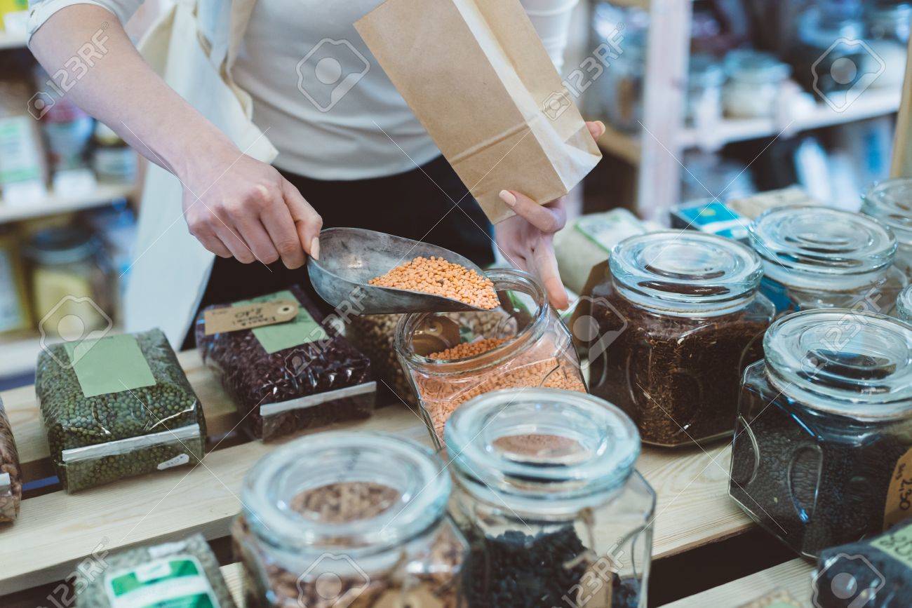 Woman puts lentil seeds into a paper bag in a healthy food store - 80017078 Woman puts lentil seeds into a paper bag in a healthy food store - 80017078