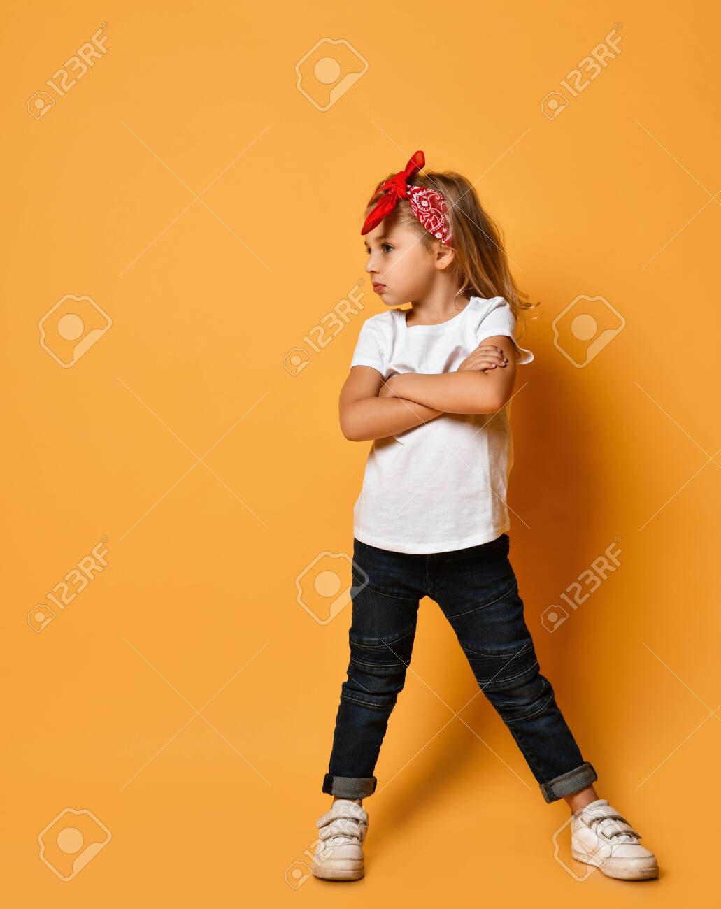 Cute Kid Blonde Girl In A White T Shirt Dark Blue Jeans And A Red Bandage On Her Head Is Standing With Her Hands Crossed Looking At Free Copy Space On Yellow Stock Cute Kid Blonde Girl In A White T Shirt Dark Blue Jeans And A Red Bandage On Her Head Is Standing With Her Hands Crossed Looking At Free Copy Space On Yellow Stock