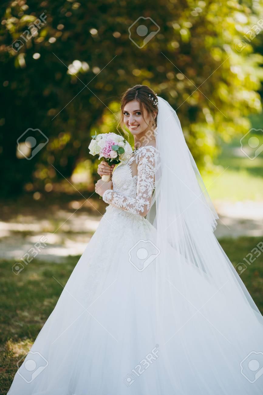 美しい結婚式のフォトセッション 風の良い晴れた日に大きな緑の庭で白とピンクの花の長いプルーム ヘアピン ベールと白とピンクの花束と白いレースのドレスを着た花嫁 の写真素材 画像素材 Image
