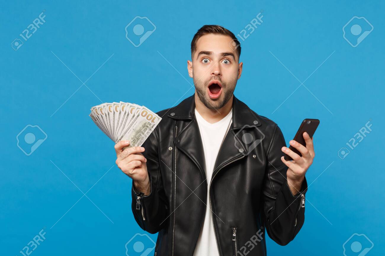 Young man in black leather jacket white t-shirt holding fan of cash money in dollar banknotes, cellphone isolated on blue wall background studio portrait. People lifestyle concept. Mock up copy space - 122091208 Young man in black leather jacket white t-shirt holding fan of cash money in dollar banknotes, cellphone isolated on blue wall background studio portrait. People lifestyle concept. Mock up copy space - 122091208