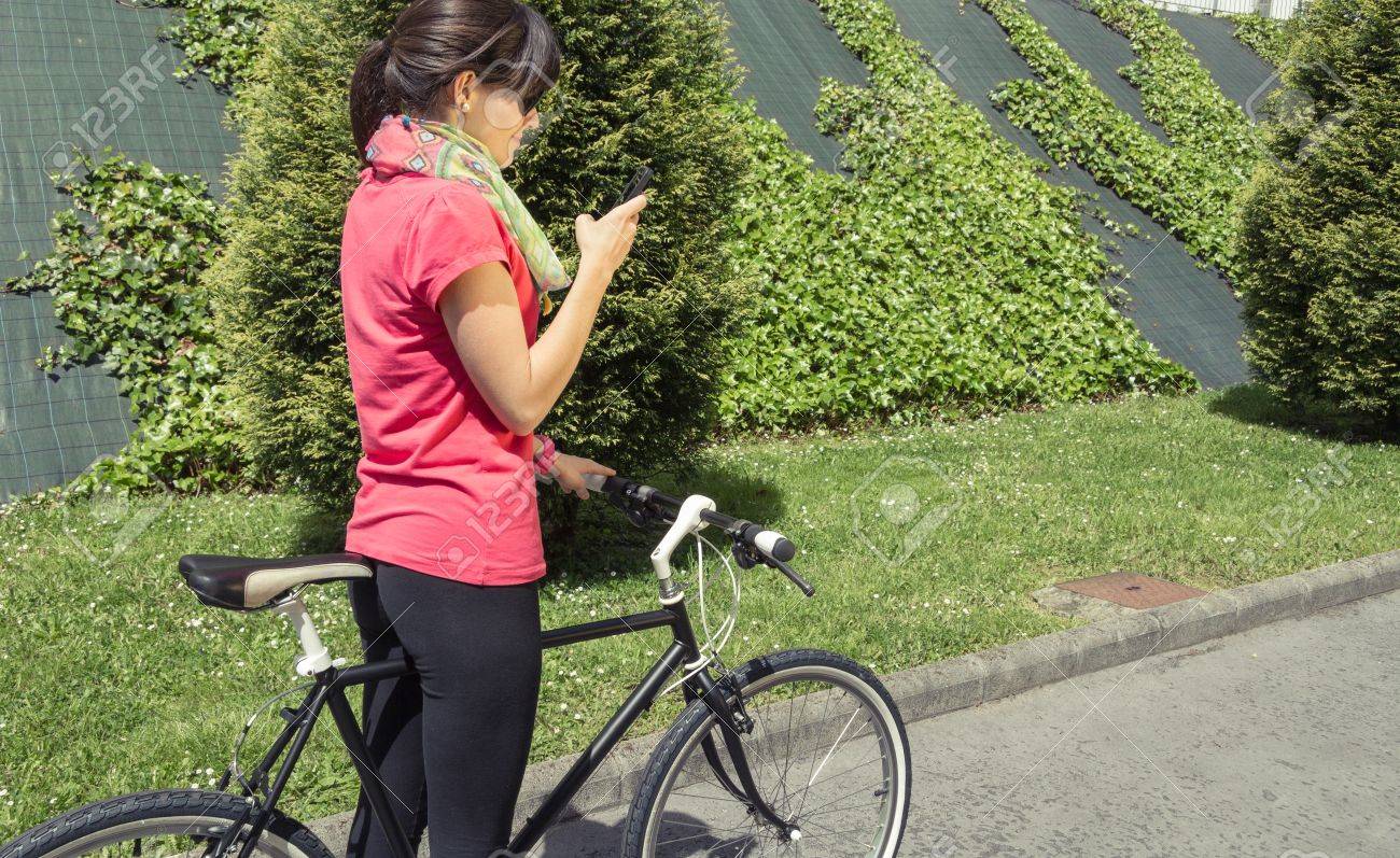 カスタム ピストと美しい若い陽気な女性のバイクのスマート フォンを探して 日当たりの良い夏の日に都市にソフトと温かみのある色調 の写真素材 画像素材 Image