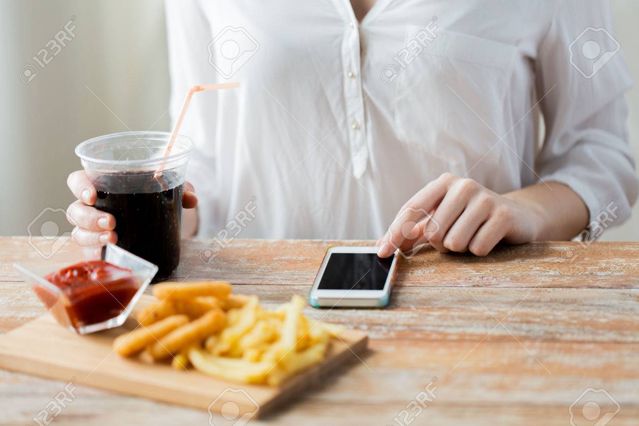 fast food, people, technology and diet concept - close up of woman with smartphone drinking coca cola and eating french fries, ketchup and deep-fried squid rings at wooden table - 54444971 fast food, people, technology and diet concept - close up of woman with smartphone drinking coca cola and eating french fries, ketchup and deep-fried squid rings at wooden table - 54444971
