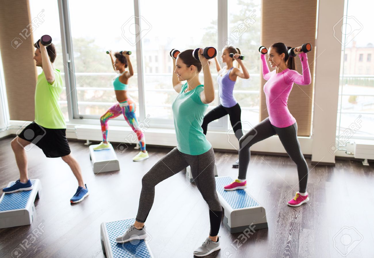fitness, sport, aerobics and people concept - group of smiling people working out with dumbbells flexing muscles on step platforms in gym - 57374799 fitness, sport, aerobics and people concept - group of smiling people working out with dumbbells flexing muscles on step platforms in gym - 57374799