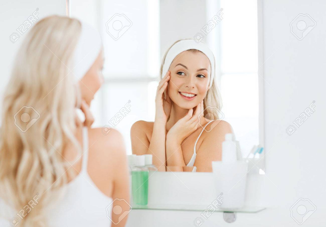 beauty, skin care and people concept - smiling young woman in hairband touching her face and looking to mirror at home bathroom - 59886820 beauty, skin care and people concept - smiling young woman in hairband touching her face and looking to mirror at home bathroom - 59886820