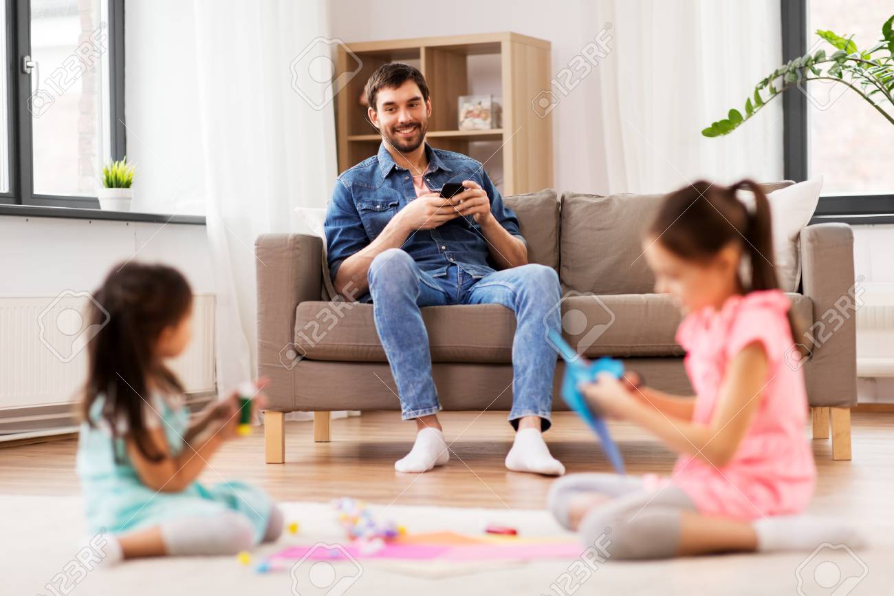 Father with smart phone looking at her daughters playing at home - 121996676 Father with smart phone looking at her daughters playing at home - 121996676