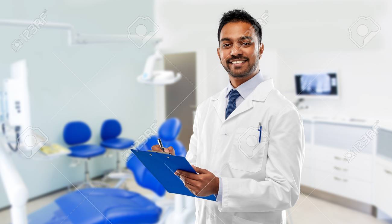 medicine, dentistry and healthcare concept - smiling indian male dentist in white coat with clipboard over dental clinic office background - 122978790 medicine, dentistry and healthcare concept - smiling indian male dentist in white coat with clipboard over dental clinic office background - 122978790