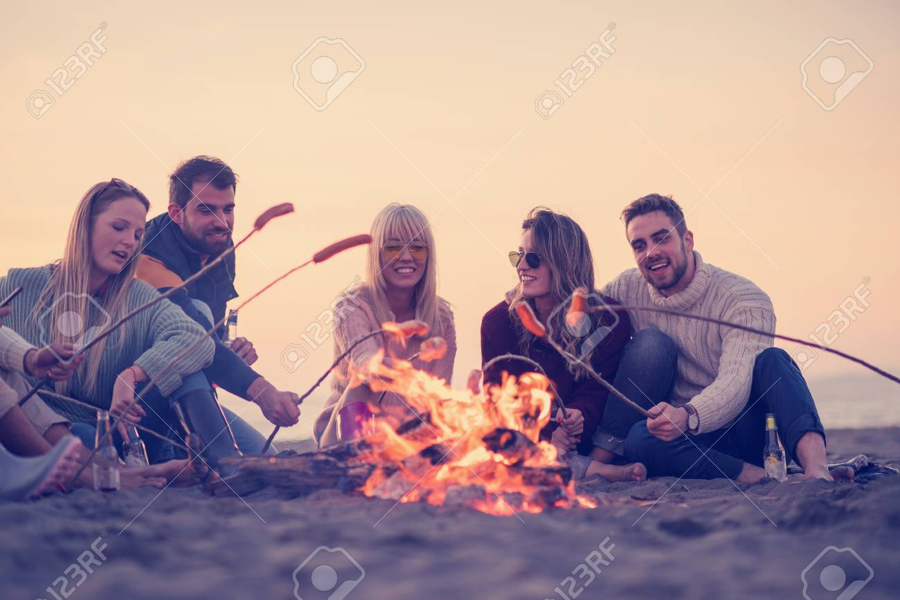 Group of young friends sitting by the fire at autumn beach, grilling sausages and drinking beer, talking and having fun - 107290336 Group of young friends sitting by the fire at autumn beach, grilling sausages and drinking beer, talking and having fun - 107290336
