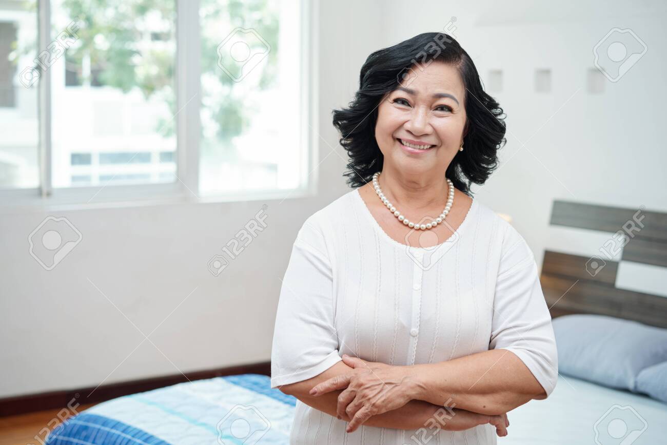 Waist-up portrait of attractive retired Asian woman with pearl necklace standing in bedroom with crossed hands and smiling at camera - 109222868 Waist-up portrait of attractive retired Asian woman with pearl necklace standing in bedroom with crossed hands and smiling at camera - 109222868