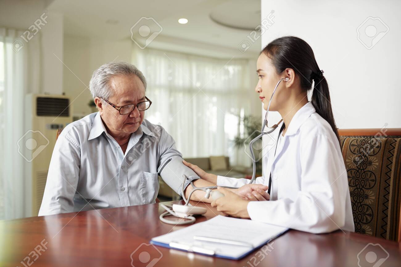 Asian young nurse sitting at the table together with elderly man and examining his blood pressure during her visit at home - 127818908 Asian young nurse sitting at the table together with elderly man and examining his blood pressure during her visit at home - 127818908