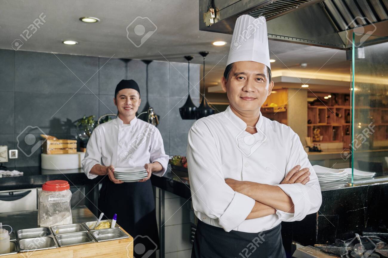 Portrait of confident Asian chef standing with arms crossed and looking at camera with young cook preparing food in the background in the kitchen - 130125469 Portrait of confident Asian chef standing with arms crossed and looking at camera with young cook preparing food in the background in the kitchen - 130125469
