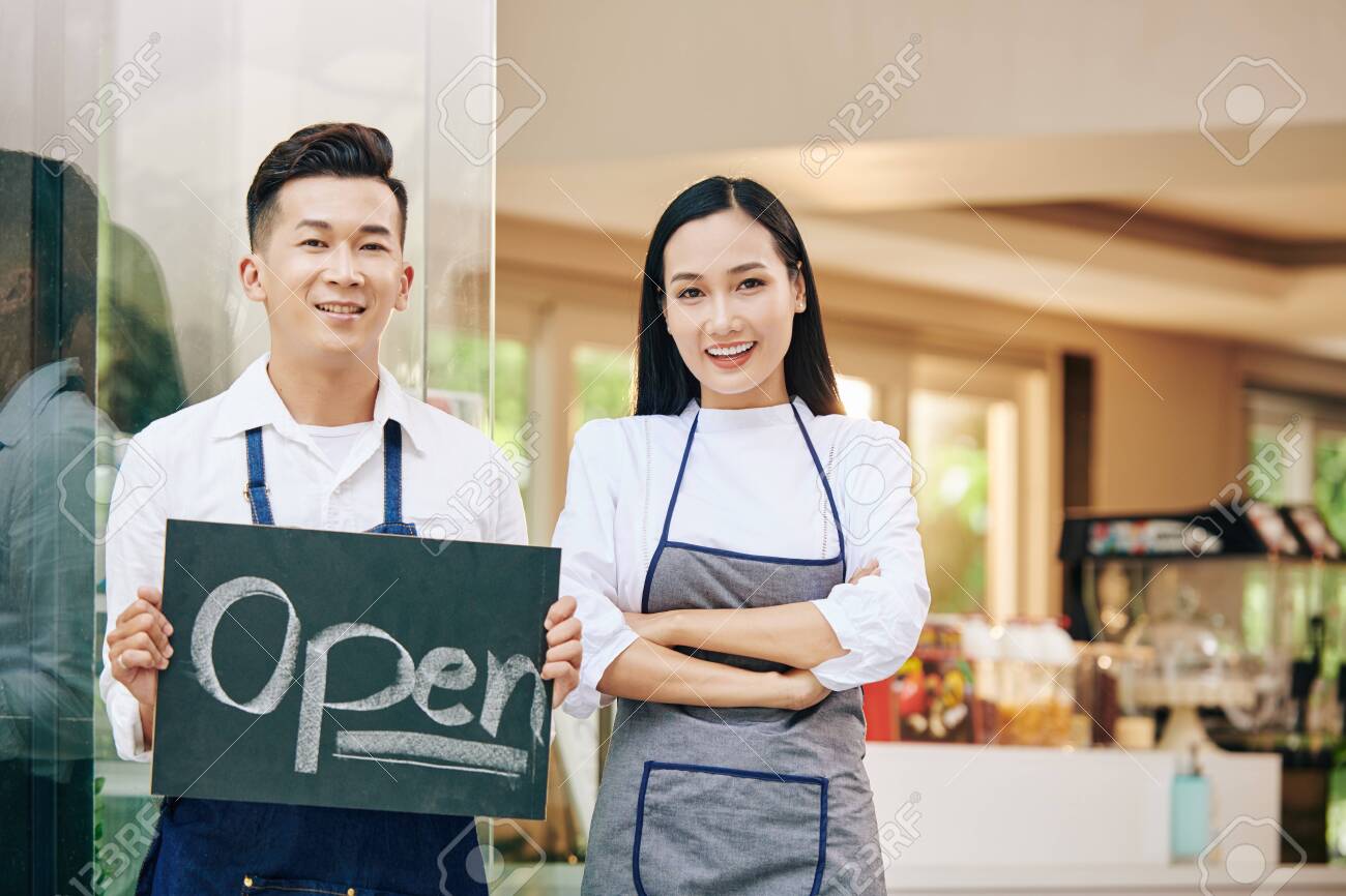 Smiling young Vietnamese cafe owners with open sign standing at cafe entrance and welcoming customers inside - 151330407 Smiling young Vietnamese cafe owners with open sign standing at cafe entrance and welcoming customers inside - 151330407