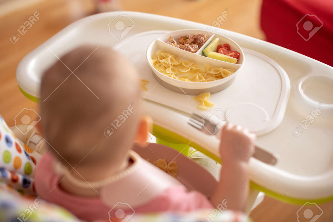 One year old girl having balanced meal in baby eating chair, healthy balanced nutrition for child, pasta, cutlet and vegetables - 166397459 One year old girl having balanced meal in baby eating chair, healthy balanced nutrition for child, pasta, cutlet and vegetables - 166397459
