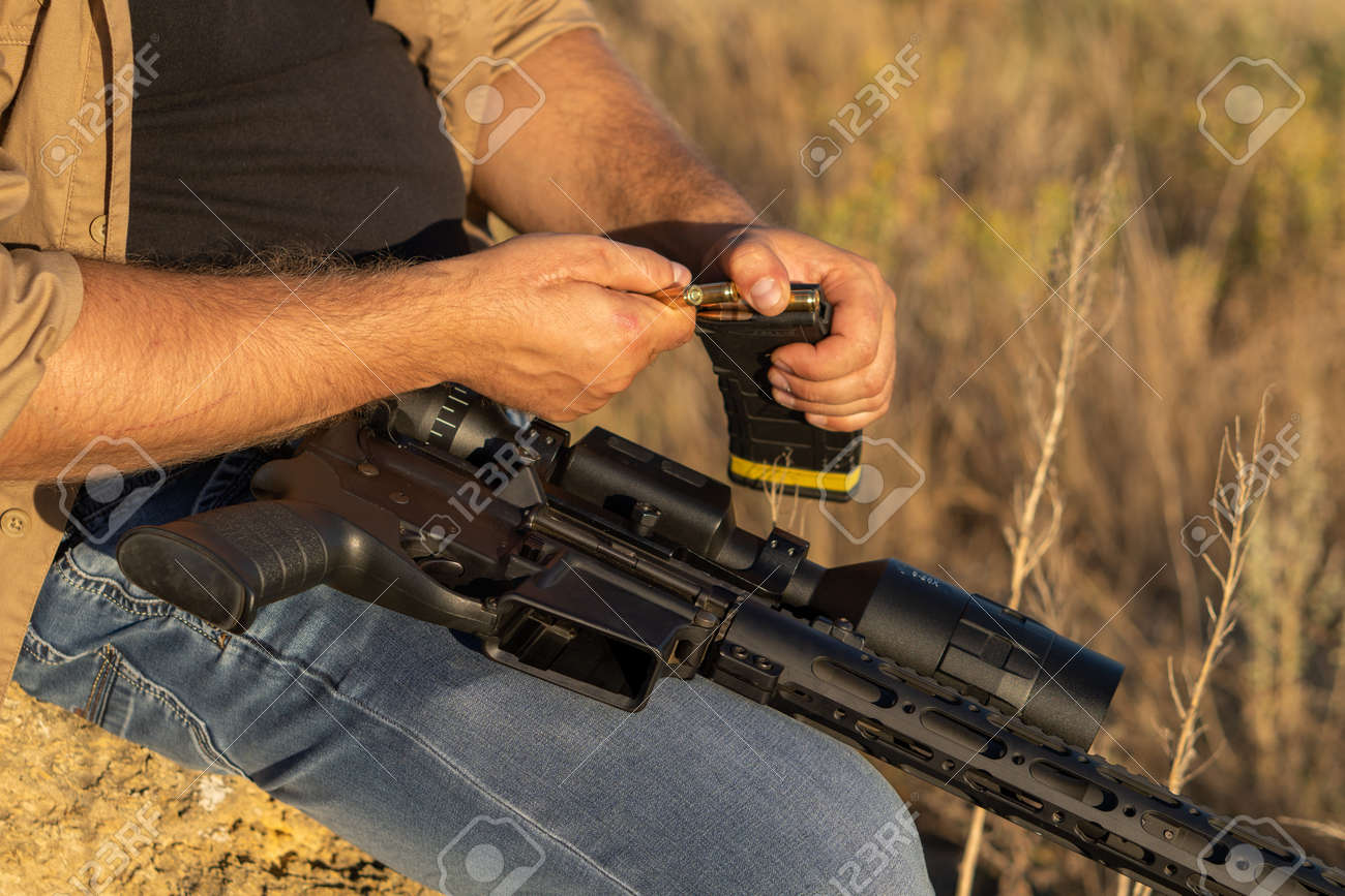 A rifle in a mans hand, close-up. Hunter with a weapon on the hunt against the background of a beautiful summer landscape - 155640479 A rifle in a mans hand, close-up. Hunter with a weapon on the hunt against the background of a beautiful summer landscape - 155640479